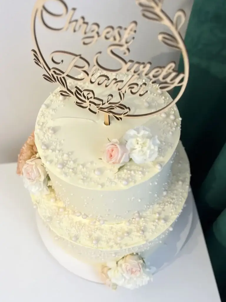 Dwupiętrowy tort przygotowany na specjalną okazje Chrztu Świętego Blanki, ozdobiony białymi i pudrowo różowymi różami z toperem wykonanmym z drewna