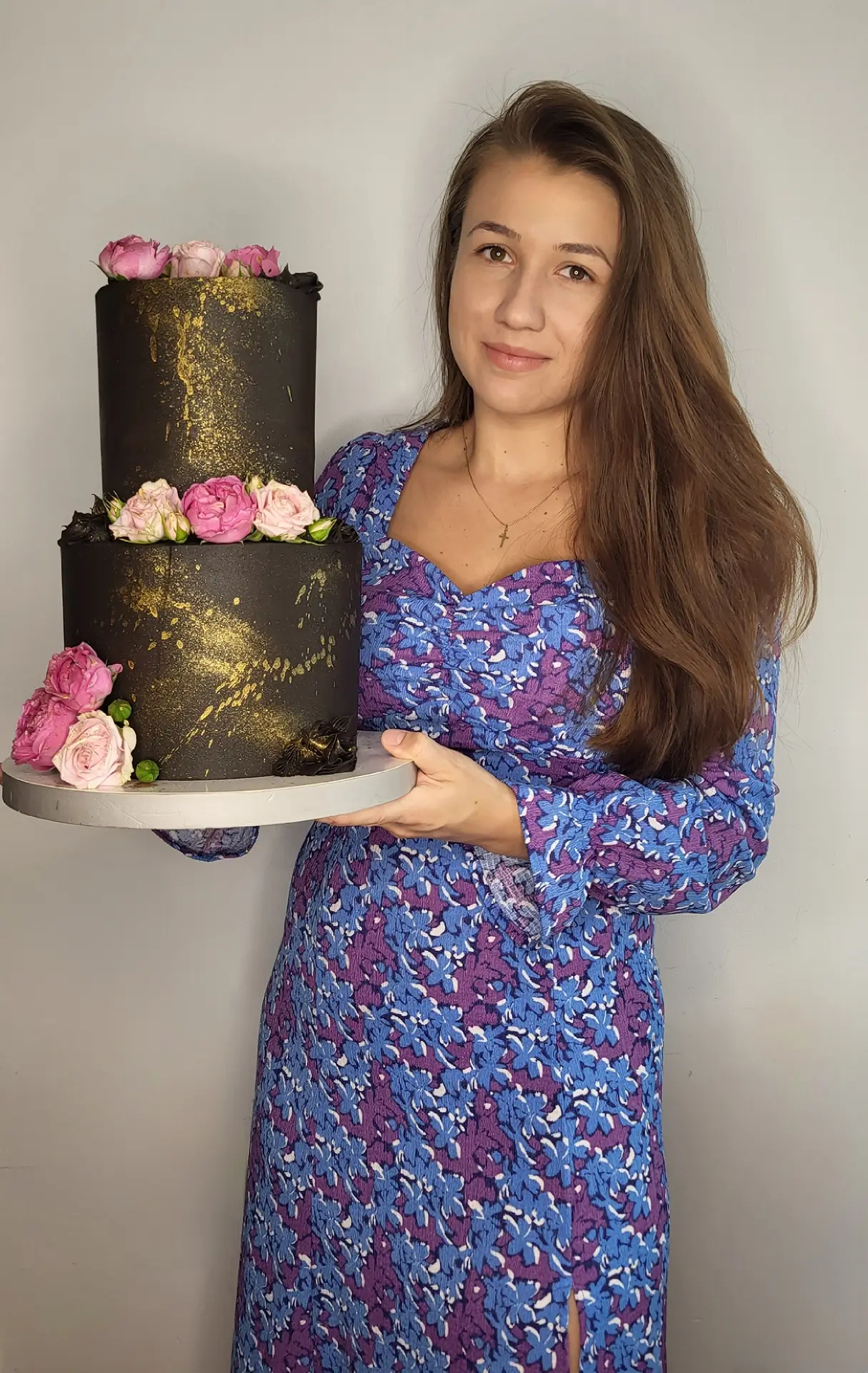 Małgorzata Poziomek, cukierniczka, trzymająca dwupiętrowy tort artystyczny z motywem czarnego tynku ze złotymi zdobieniami i różowymi kwiatami, ubrana w niebieską sukienkę w kwiaty