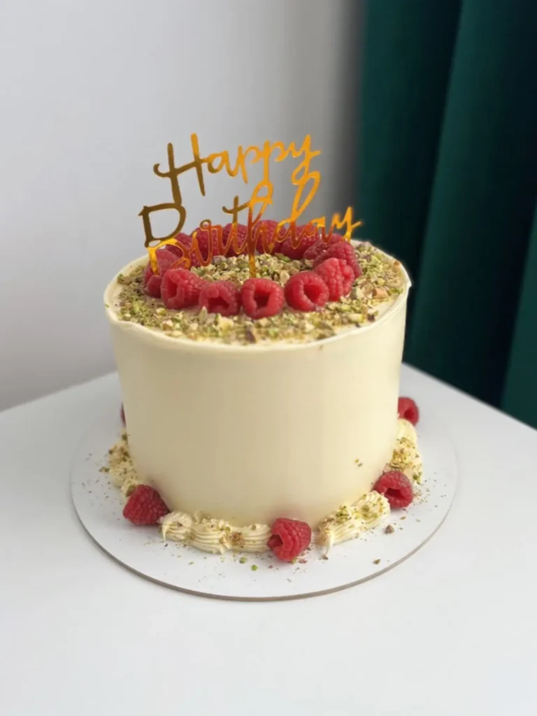 Klasyczny tort urodzinowy ozdobiony świeżymi malinami posypany świeżymi pistacjami z złotym toperem