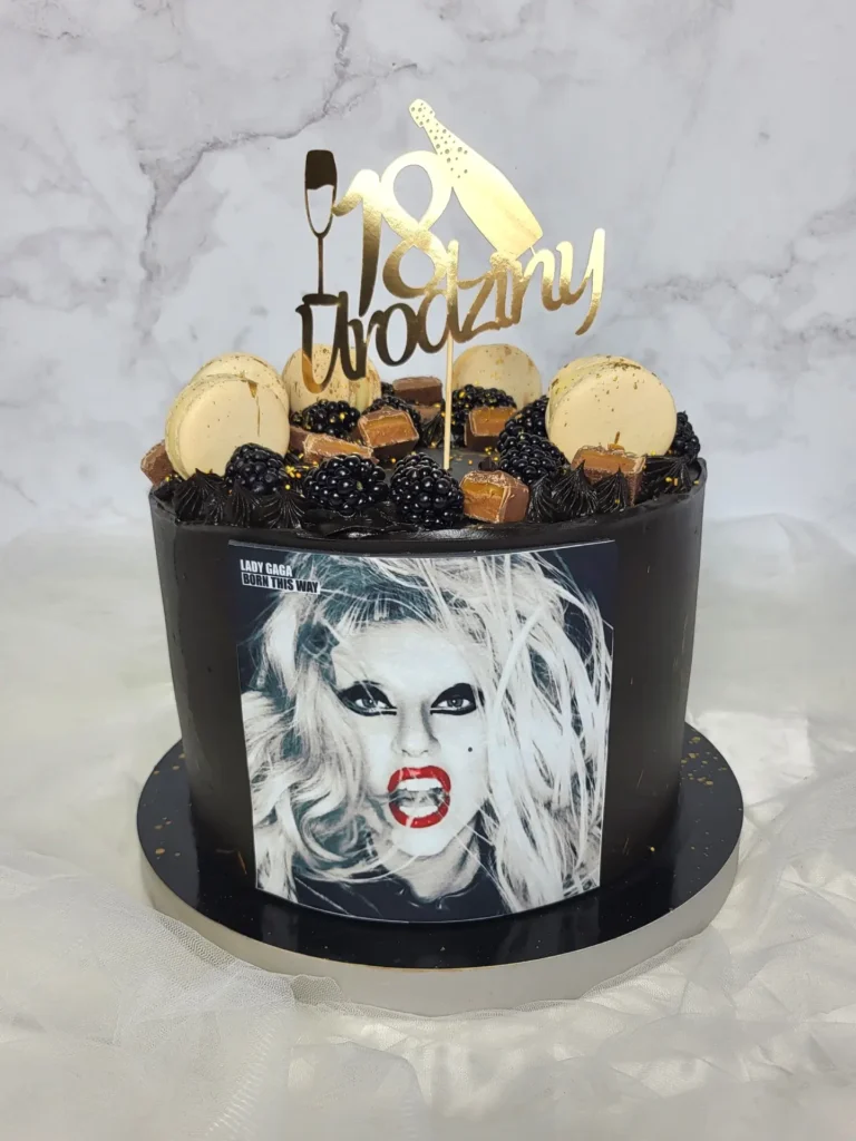 Tort na osiemnaste urodziny z czarnym tynkiem. Przyozdobiony złotym toperem, kremowymi makaronikami i czarnymi owocami oraz zdjęciem piosenkarki "LADY GAGA"